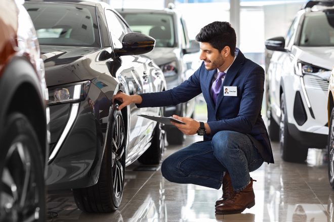 Handsome man in suit sales assistant checking car tires at luxury showroom, holding folder with papers. Middle-eastern guy sales manager in auto salon making checkup, touching automobile wheel